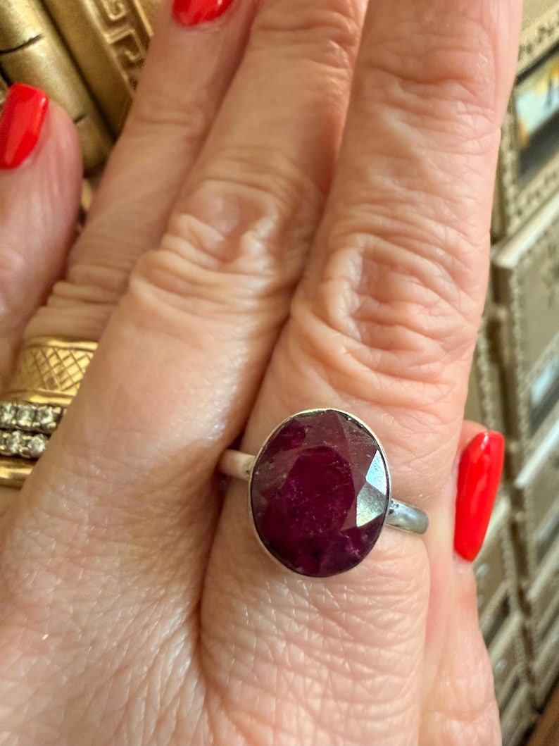 May include: A silver ring featuring a large, oval-cut ruby gemstone. The ruby is a deep red color and set in a simple silver band. The ring is shown on a hand with red nail polish.