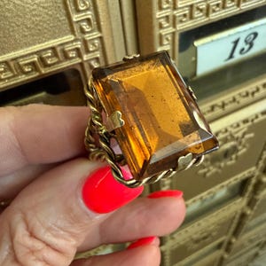 May include: A vintage gold-toned ring featuring a large, rectangular amber-colored gemstone. The gemstone is set in a twisted rope-like band with heart-shaped accents. The ring is a statement piece, showcasing a bold and classic design.