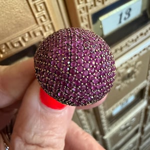 May include: A dome-shaped ring featuring a dense cluster of small, round, deep pink gemstones set in a metallic base. The surface of the ring is entirely covered in these faceted stones, creating a textured, spherical appearance on the top portion.