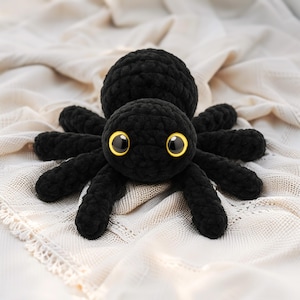 Puede incluir: Araña negra de ganchillo hecha a mano con ocho patas y grandes ojos con borde amarillo. El peluche está sobre una tela crema texturizada, creando un suave contraste. Un artículo decorativo único y caprichoso.