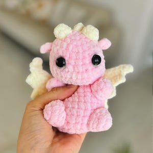 May include: A handmade pink crochet dragon with large black eyes and small cream-colored horns and wings. The plush toy is soft and cuddly, perfect for a child's bedroom or nursery. The dragon is a whimsical and charming addition to any collection.