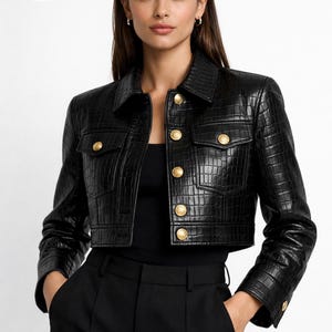 May include: Black cropped leather jacket with a crocodile texture, featuring gold buttons and two front pockets. The jacket has a classic collar and long sleeves. The brand logo "PLANIO LEATHER" is in the top left corner.