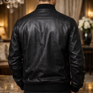K&ouml;nnte beinhalten: Schwarze Leder-Bomberjacke mit geripptem Kragen und B&uuml;ndchen. Die Jacke hat ein klassisches Design mit glatter, gl&auml;nzender Oberfl&auml;che. Das Model tr&auml;gt dunkle Jeans. Der Hintergrund ist eine verschwommene Innenaufnahme.