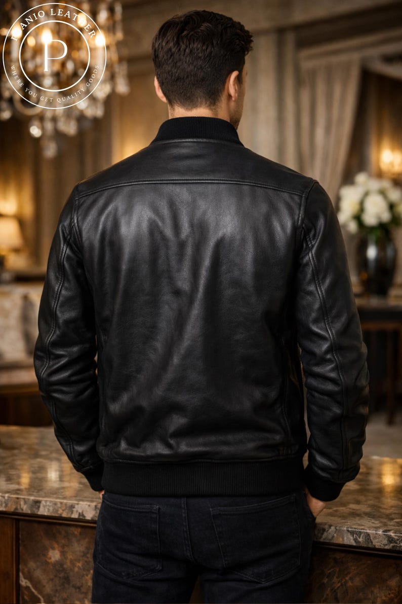 K&ouml;nnte beinhalten: Schwarze Leder-Bomberjacke mit geripptem Kragen und B&uuml;ndchen. Die Jacke hat ein klassisches Design mit glatter, gl&auml;nzender Oberfl&auml;che. Das Model tr&auml;gt dunkle Jeans. Der Hintergrund ist eine verschwommene Innenaufnahme.