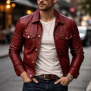 May include: A deep red leather jacket with a classic collar and buttoned chest pockets. The jacket is worn over a white t-shirt and paired with dark blue jeans. A brown leather belt and a silver watch complete the outfit. The image features the "PLANIO LEATHER" logo.