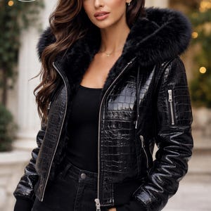 May include: Black leather jacket with a faux fur-lined hood. The jacket has a glossy, textured finish and a zipper closure. The sleeves have ribbed cuffs. The jacket is worn with black jeans and a black top. The logo "PLANIO LEATHER" is visible.