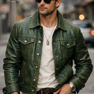 Könnte beinhalten: Grüne Lederjacke mit Used-Look, zwei Brusttaschen und Knopfverschlüssen. Sie wird über einem weißen T-Shirt und mit dunkelblauen Jeans getragen. Die Abbildung zeigt auch eine silberne Uhr und einen braunen Ledergürtel.