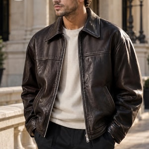 May include: A dark brown leather jacket with a classic collar, a full-length silver metal zipper, and two side pockets. The jacket is worn over a cream-colored knit sweater and black trousers. A circular logo in the corner reads PLANIO LEATHER.