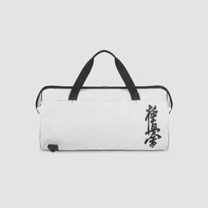 Puede incluir: Una bolsa de deporte blanca con asas negras y cierre de cremallera negro. La bolsa presenta un símbolo kanji caligráfico negro impreso verticalmente en el lado derecho. El diseño incluye un pequeño punto de sujeción negro en la esquina inferior izquierda.