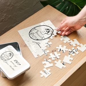 May include: A white jigsaw puzzle featuring a black and white portrait of Jane Austen, with the text "250 years of Jane Austen." The puzzle is partially assembled, with pieces scattered on a wooden surface. A metal tin with the same image is visible.