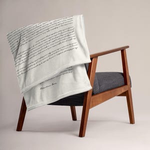 Könnte beinhalten: Eine weiße Wurkdecke mit schwarzem Schrifttext, die über einem Holzstuhl mit grauem Sitz drapiert ist. Die Decke weist einen handschriftlichen Textstil auf. Der Stuhl hat ein modernes Design aus der Mitte des Jahrhunderts.