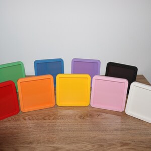 May include: A selection of square trays in a range of colours: red, orange, yellow, pink, green, blue, purple, black, and white. The trays are displayed on a wooden surface, highlighting their bright colours and simple design.