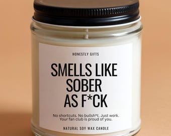 May include: A clear glass jar candle with a black lid. The candle has a white label with the text "SMELLS LIKE SOBER AS F*CK". Below the text are the words "No shortcuts. No bullsh*t. Just work. Your fan club is proud of you."