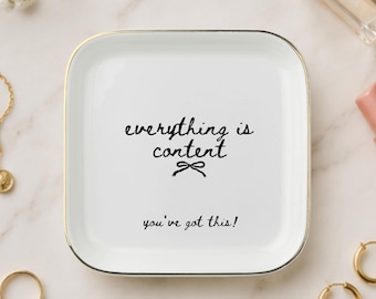 May include: A square white ceramic trinket dish with rounded corners and a gold-toned rim. The center features black cursive text that reads everything is content with a small bow icon below it, and the phrase you've got this! at the bottom.