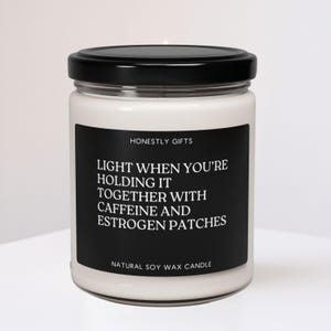 May include: A white soy wax candle in a clear glass jar with a black lid. The candle has a black label with the text "LIGHT WHEN YOU'RE HOLDING IT TOGETHER WITH CAFFEINE AND ESTROGEN PATCHES."