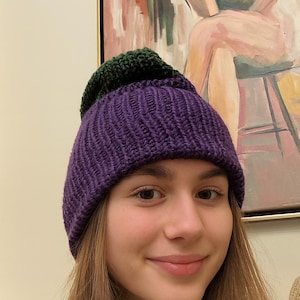 May include: A knitted beanie hat in two colors: a deep purple base and a dark green top. The hat is made of a thick, textured yarn. The hat is worn by a person.