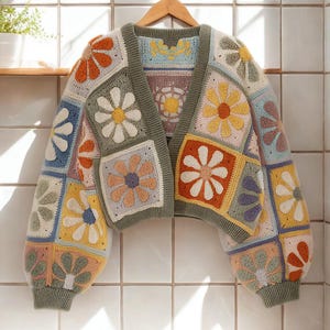 May include: A colorful, crocheted cardigan with a daisy flower pattern. The sweater features a patchwork design with various pastel colors, including orange, yellow, blue, and green. The sleeves are puffy, and the cardigan is displayed on a wooden hanger.