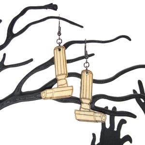 May include: Two wooden earrings in the shape of a vintage vacuum cleaner. The earrings are light brown with etched details and hang from dark metal hooks. The earrings are displayed against a black, stylized tree and hand silhouette.