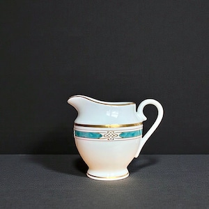 May include: A white ceramic creamer with a turquoise and gold band design. The creamer has a curved spout and a small handle. The base and rim are trimmed with gold. The creamer is approximately 4 inches tall.