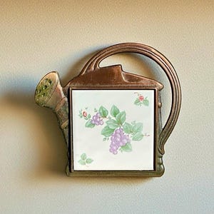 May include: A decorative watering can-shaped wall hanging. The metal frame has a bronze finish, and the center features a square tile with a floral design of purple grapes and green leaves. The spout has a green, textured finish.