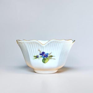 May include: A white porcelain bowl with a scalloped rim and vertical ribbed texture. The bowl features a hand-painted floral design with blue flowers, green leaves, and gold trim. The bowl is a decorative piece.