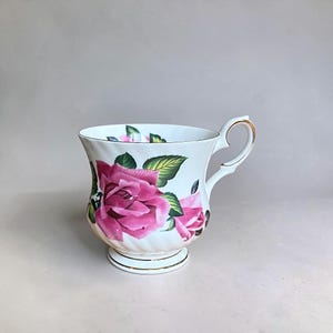 May include: White teacup with a pink rose floral design and gold trim. The cup has a curved handle and a pedestal base. Green leaves accent the pink roses. A classic teacup for serving tea.