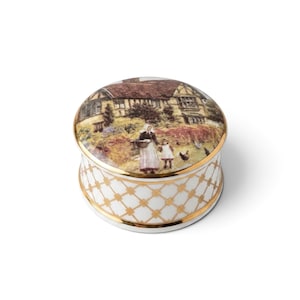 May include: A round porcelain trinket box with a gold-trimmed lid featuring a colorful illustration of a cottage, a person, and a child in a garden. The base is white with a gold lattice pattern and gold bands at the top and bottom edges.