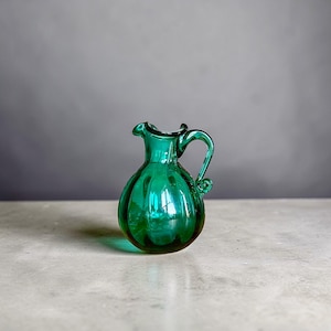 May include: A small, emerald green glass pitcher with a rounded body and a handle. The pitcher has a ruffled rim and vertical ridges. The handle has a decorative swirl at the base. The pitcher is sitting on a light-colored surface.