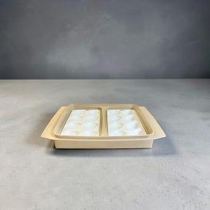 May include: A beige plastic paint palette with two sections, each containing multiple wells for holding paint. The palette sits inside a larger, matching tray. The wells are filled with a creamy white substance.