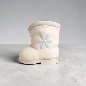 May include: A cream-colored ceramic boot-shaped planter featuring a light blue daisy flower with a yellow center on the side. The top rim of the boot has a decorative embossed stitch pattern in a light blue color, providing a subtle contrast.
