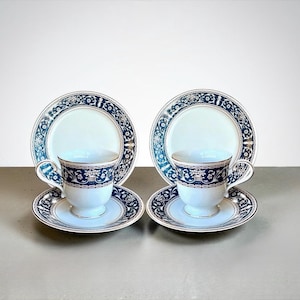 May include: Two matching teacups, saucers, and plates. The white porcelain cups and saucers have a navy blue floral design with gold trim. The plates have a similar navy blue floral design around the rim. The set is displayed on a neutral surface.