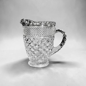 May include: Clear glass pitcher with a diamond pattern and a decorative rim. The pitcher has a handle and a spout for pouring. The glass is transparent, allowing light to pass through. The pitcher is likely used for serving beverages.