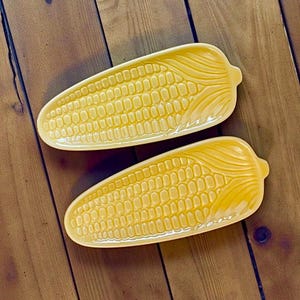 May include: Two yellow ceramic corn-shaped serving dishes. The dishes are detailed with rows of kernels and a husk design at the end. They are placed on a wooden surface.