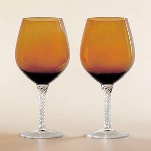 May include: Two amber-colored wine glasses with a dark base and a clear, twisted stem. The glasses have a classic tulip shape and sit on a clear, round base. The background is a neutral beige.
