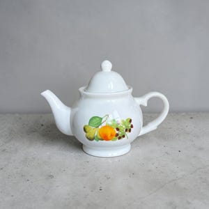 May include: White ceramic teapot with a lid and a handle. The teapot features a colorful fruit design with an orange, green leaves, and red berries. The teapot is on a gray surface.