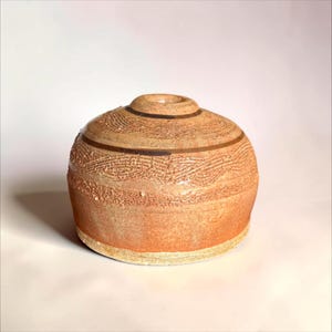 May include: A round, low-profile ceramic vessel with a small central opening. The exterior features a textured, reddish-brown clay surface with horizontal bands of intricate, raised wave-like patterns and smooth dark brown lines.