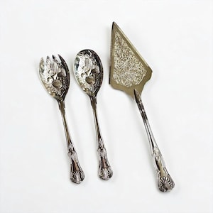 May include: A set of three silver-toned metal utensils including a salad fork, a spoon, and a triangular cake server. Each piece features an ornate, embossed design on the handle and a detailed, intricate pattern on the bowl or blade surface.