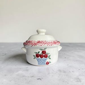 May include: A small, white ceramic casserole dish with a lid. The rim of the lid and the top of the dish are decorated with a red and white pattern. The front of the dish features a blue basket filled with red apples.