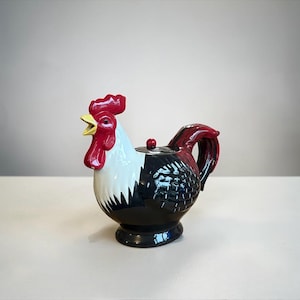 May include: A ceramic teapot shaped like a rooster. The teapot features a red comb, wattle, and handle, with a white and black body. The beak is yellow, and the teapot sits on a black base.