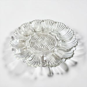 May include: A clear glass oyster plate featuring a central circular section with a radiating sunburst pattern, surrounded by twelve individual shell-shaped indentations arranged in a ring, each with fine vertical ridges and a smooth scalloped edge.