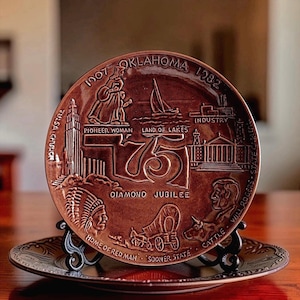 May include: A round brown commemorative plate celebrating the Oklahoma Diamond Jubilee 1907-1982. The plate features embossed imagery including a pioneer woman, a sailboat, a covered wagon, a Native American figure, and text detailing Oklahoma history.