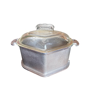 May include: A vintage, square-shaped casserole dish with a clear glass lid. The dish has a textured, silver-colored metal base and handles. The glass lid features a decorative pattern and a handle. A classic kitchenware item.