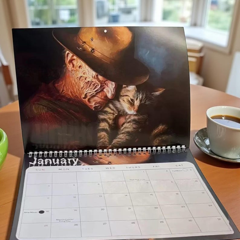 May include: A January calendar featuring a portrait of a character from a horror film holding a tabby cat. The calendar is open, with a cup of coffee on a saucer to the right. The calendar is on a wooden table.