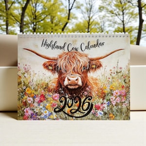 May include: A spiral-bound calendar featuring a watercolor illustration of a Highland cow. The cow has long, shaggy, reddish-brown fur and large horns. The calendar is titled "Highland Cow Calendar" with the year "2026" in a decorative script.