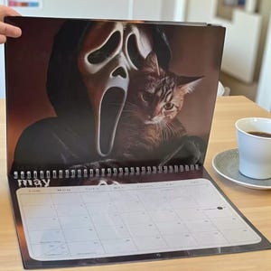 May include: A spiral-bound calendar open to the month of May. The calendar features a spooky image of a cat and a Scream mask. A cup of coffee sits on a saucer in the background.