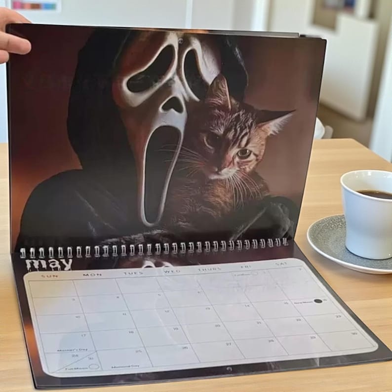 May include: A spiral-bound calendar open to the month of May. The calendar features a spooky image of a cat and a Scream mask. A cup of coffee sits on a saucer in the background.