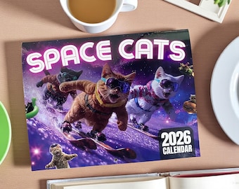 May include: A 2026 calendar titled "SPACE CATS" features cats wearing goggles and riding snowboards on a purple and blue background. The calendar is open on a wooden surface, with a cup of coffee and a book visible.