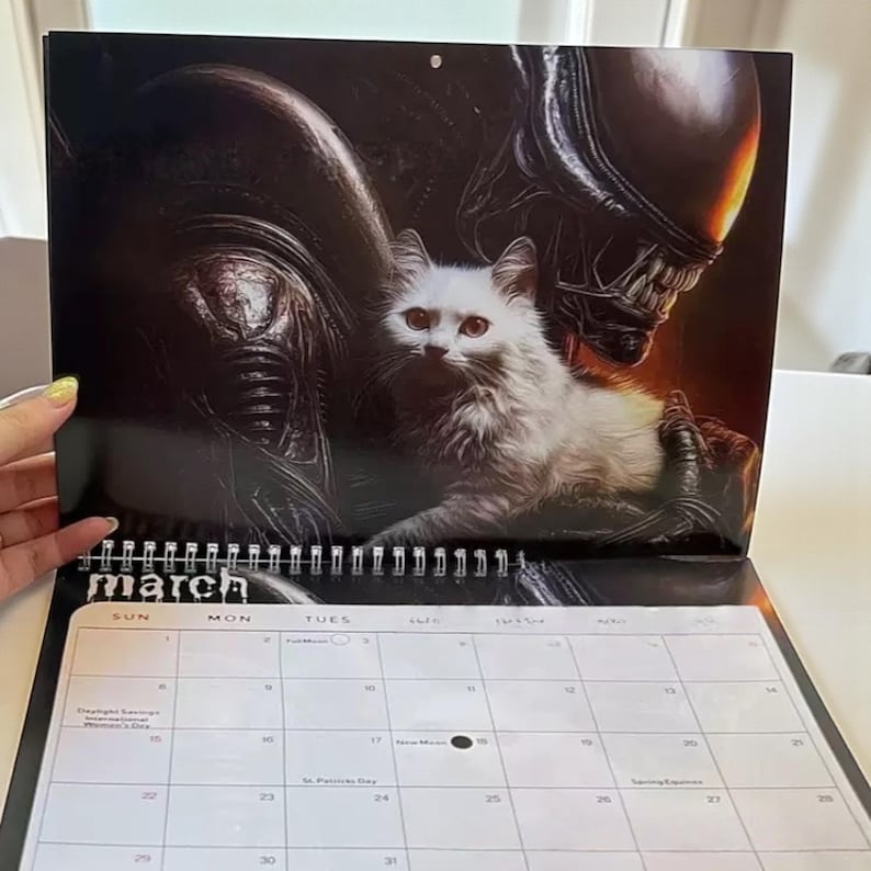 May include: A March calendar featuring a white fluffy cat held by an Alien creature. The calendar has a spiral binding and a white background with the days of the month. The Alien is dark with a menacing appearance.