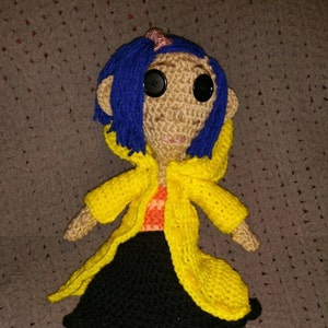 May include: Crocheted doll featuring a tan face with large black button eyes, blue hair, and a yellow coat. The doll is wearing an orange shirt and a black skirt. A pink hair accessory is visible.