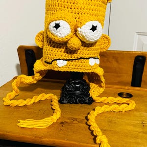 May include: A handmade, yellow crochet hat resembling a cartoon character's head. It has large white eyes with black star pupils, a small nose, and two white teeth. Long, braided yellow tassels hang from the sides of the hat.